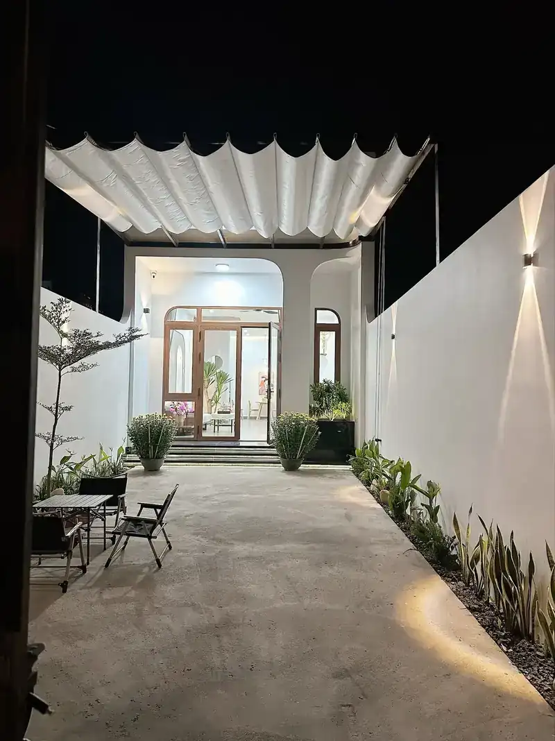 Sân trước B-House nhà cấp 4 5x20 Kon Tum đêm — mái bạt xếp, cửa vòm gỗ, tường trắng