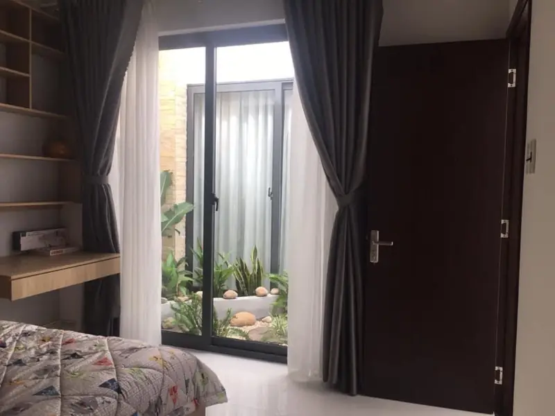 Phòng ngủ nhà cấp 4 nhìn sân trong — cửa kính, rèm xám, bàn gỗ, HQ-House Kon Tum